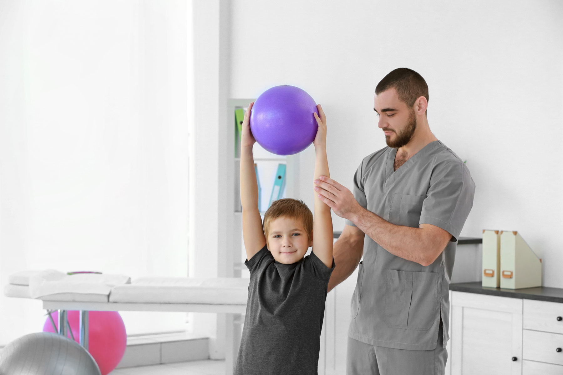 Physical therapist helping a boy lift a ball overhead during pediatric physical therapy in El Paso